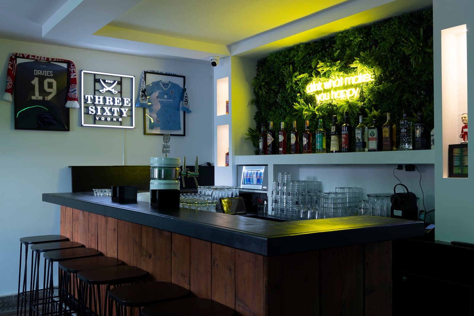 Tresen der FIFTY57SEVEN Bar Dachau mit Zapfanlage, Spirituosen vor der Pflanzenwand und gerahmten Fußballtrikots an der Wand