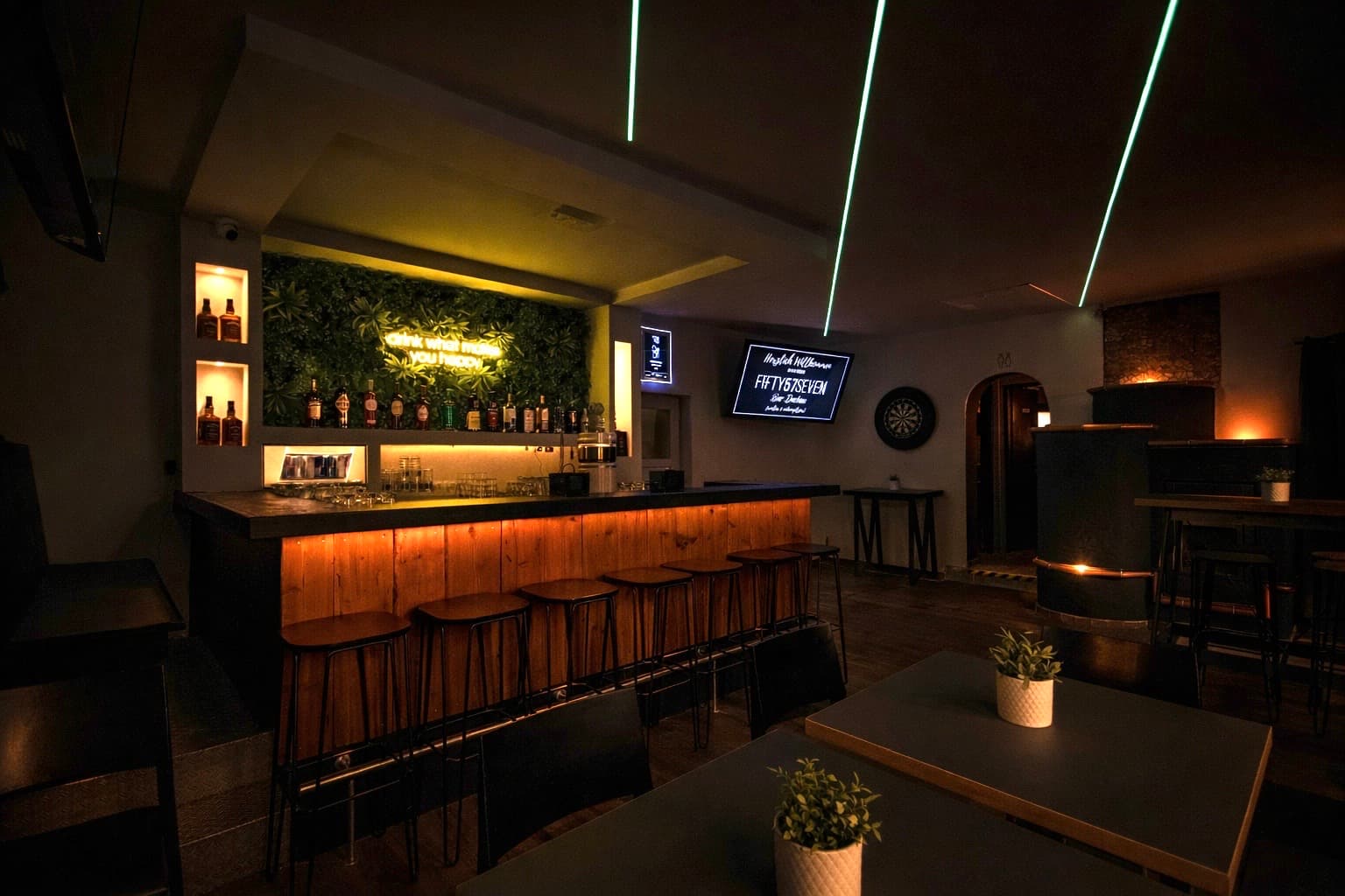 Innenraum der FIFTY57SEVEN Bar Dachau am Abend: beleuchteter Holztresen, Barhocker, Pflanzenwand mit Neonschriftzug und grüne Laserlinien an der Decke
